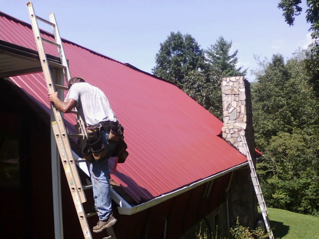 Roofer on a ladder beside a red metal roof and stone chimney — Soffit & Fascia Repair by Roofing Brothers in Wharton